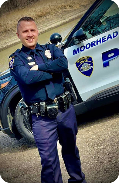 Brad Schenck in his police uniform smiling in front of a Moorhead Police Department vehicle.